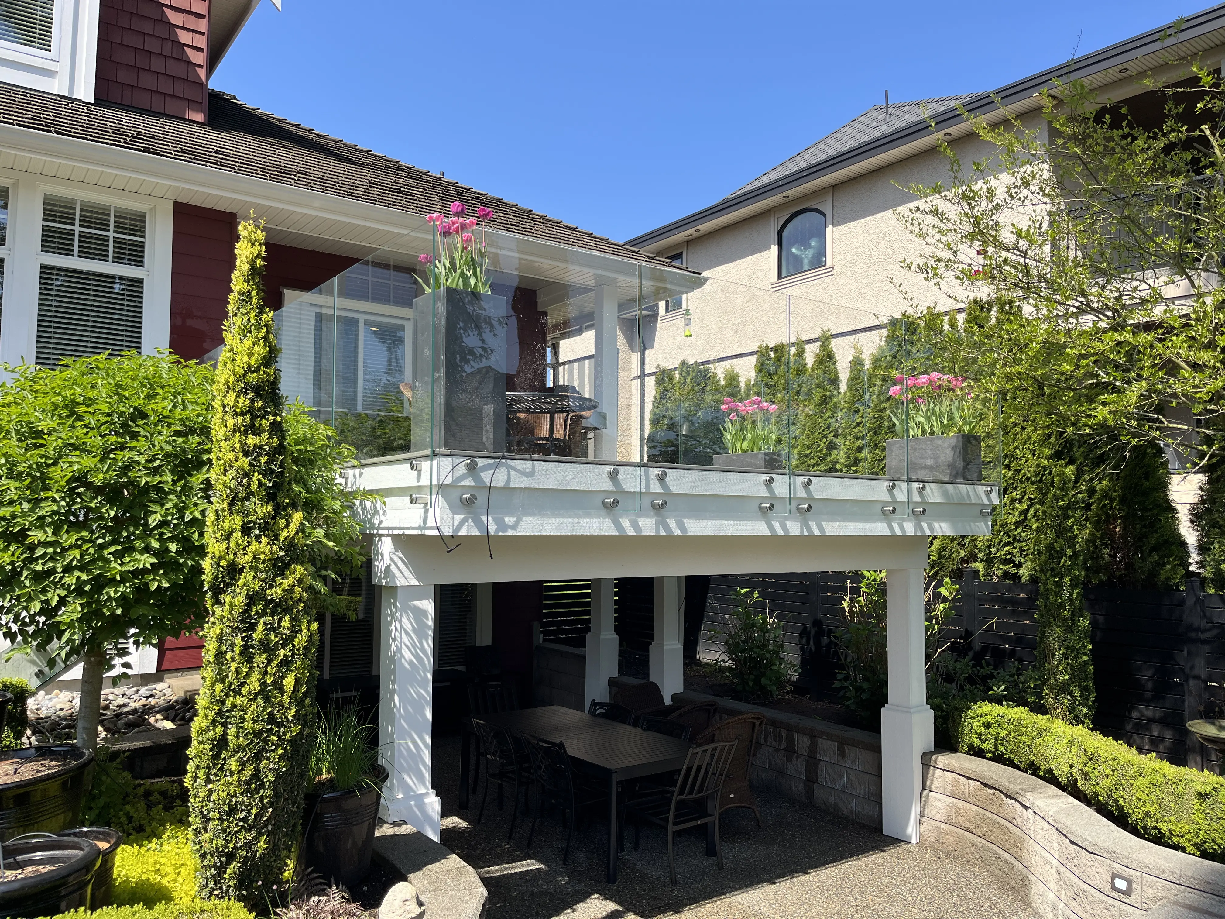 Elevated backyard deck with frameless glass railing above a covered outdoor dining area in a landscaped residential yard.