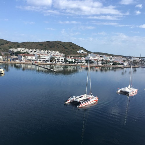 Två katamaraner ankrade i en lugn hamn med en stad och kullar i bakgrunden under en delvis molnig himmel.