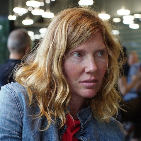 Architect and author Christie Pearson, with long wavy reddish-blond hair and a denim jacket over a red top, looks to the side at an event, people and lights blurred behind her.