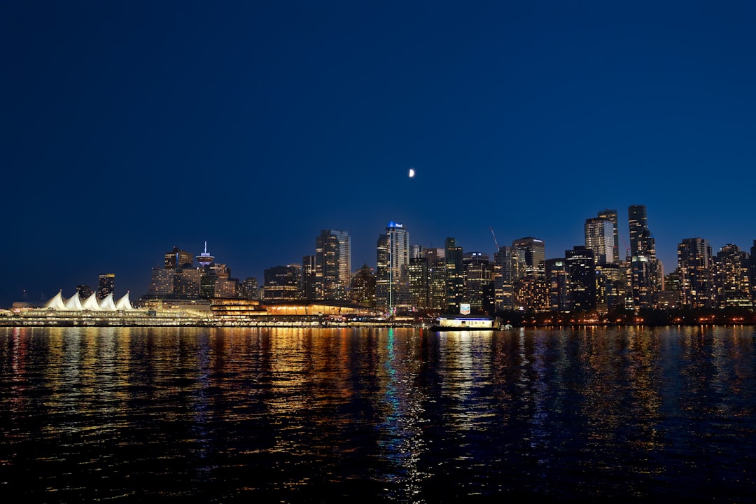 City skyline reflected in calm harbor water at night with a crescent moon above.