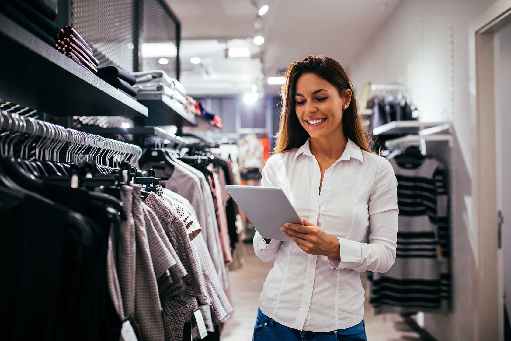A smiling woman in a white shirt using a tablet in a clothing store, standing near racks of garments to manage inventory.