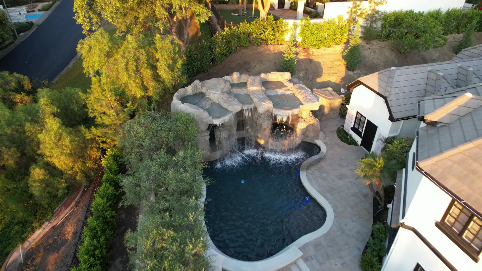 Aerial view of a curved swimming pool, rock waterfall, paved deck and surrounding planting.