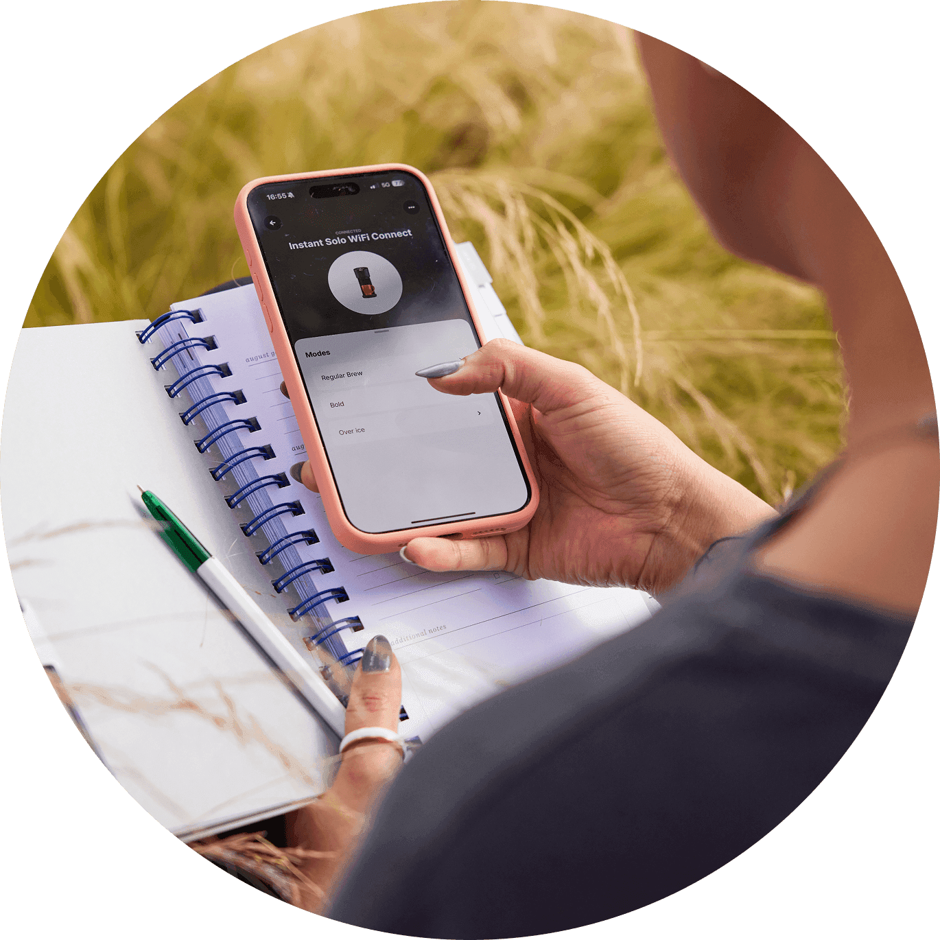 Person holds a smartphone showing a connected cooking app while sitting outdoors with a notebook and pen