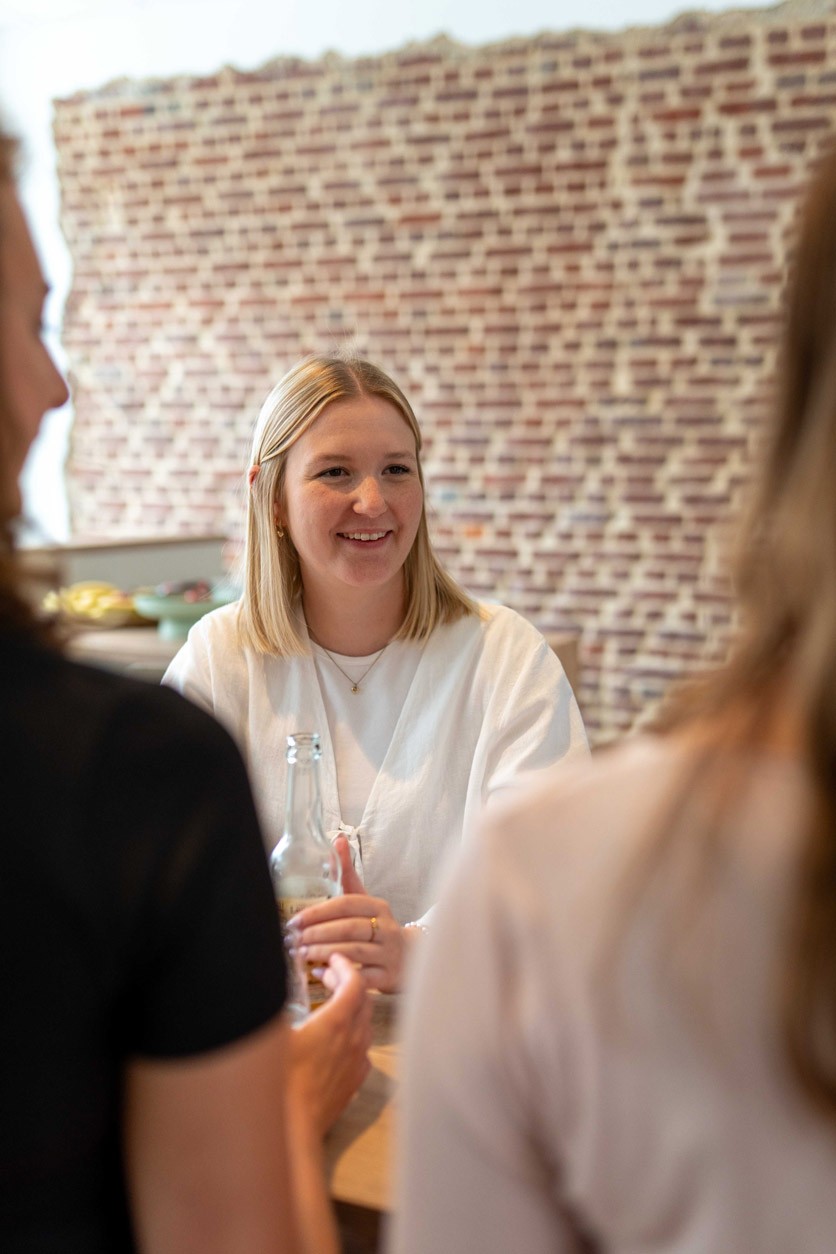 Mitarbeiterin im hellen Oberteil sitzt im Gespräch an einem Tisch, im Hintergrund eine Backsteinwand.