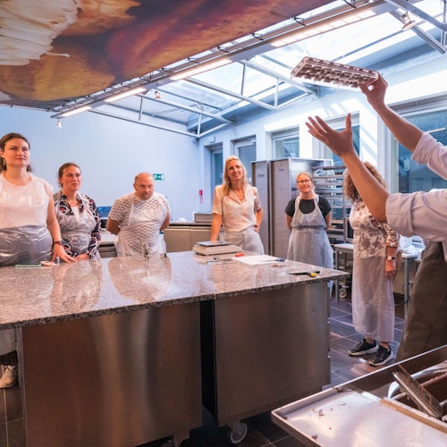 Egy csoport kötényes ember figyelmesen nézi, ahogy egy oktató egy főzési technikát bemutat egy modern konyhában.