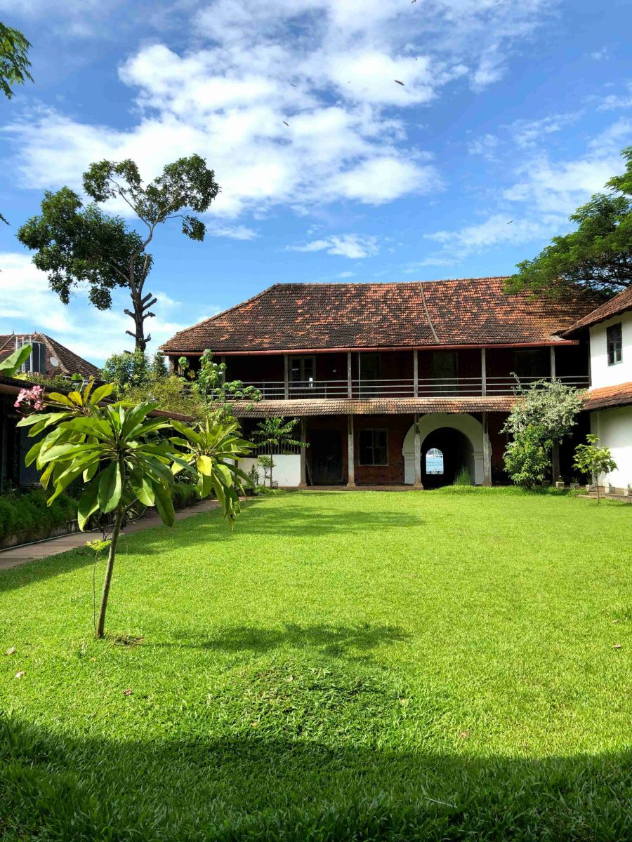 Two-story building with a terracotta tiled roof and archway overlooking a lush green lawn under a sunny sky with clouds.