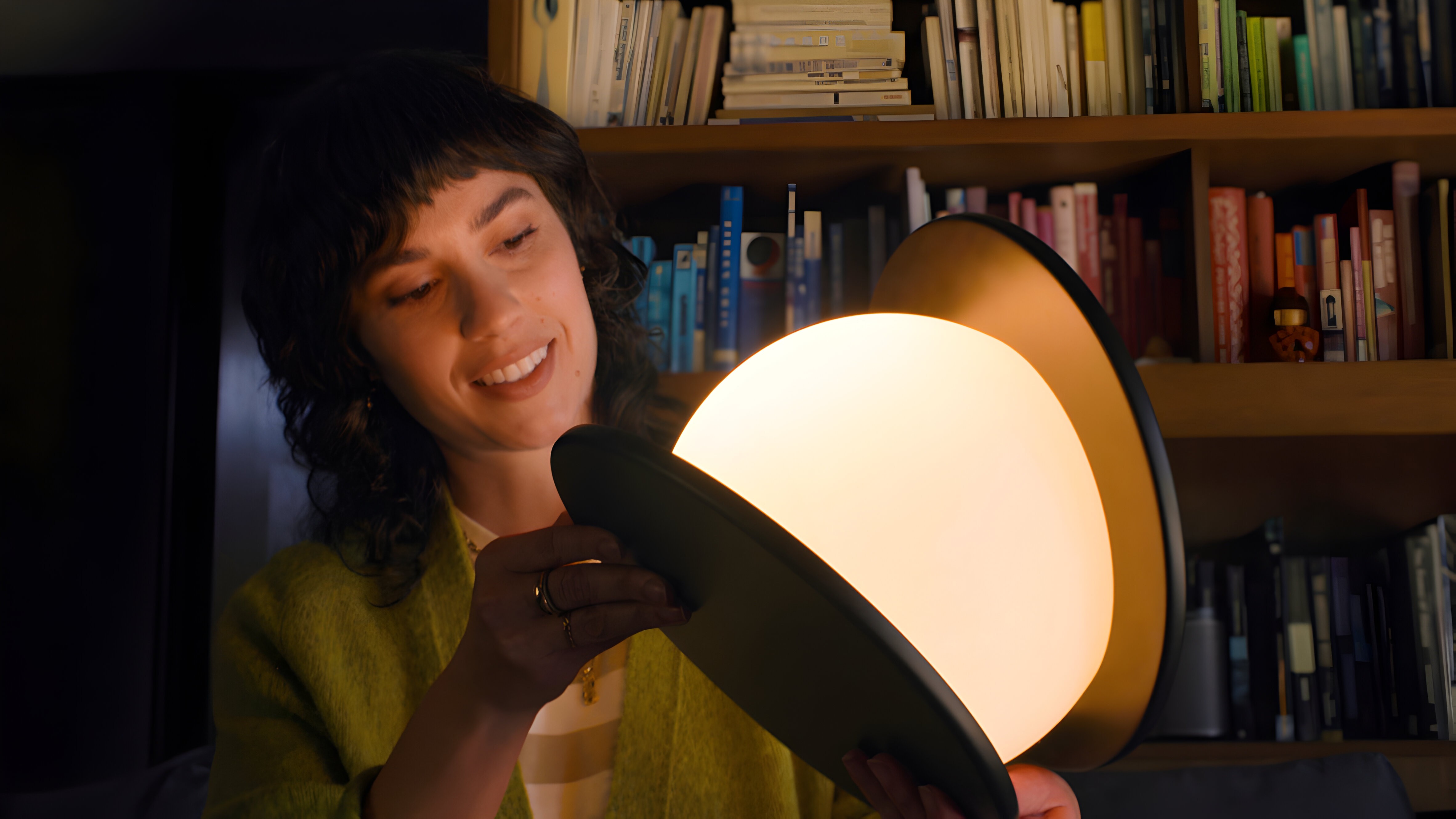 Smiling woman holding a pendant light's two discs apart around its glowing globe, in front of a bookshelf