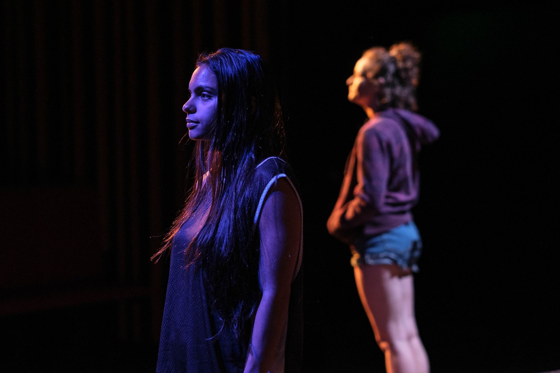 Two performers stand apart on a dim stage: one in the foreground gazes ahead in blue light, while the other stands behind her in soft focus, creating a sense of distance and tension.