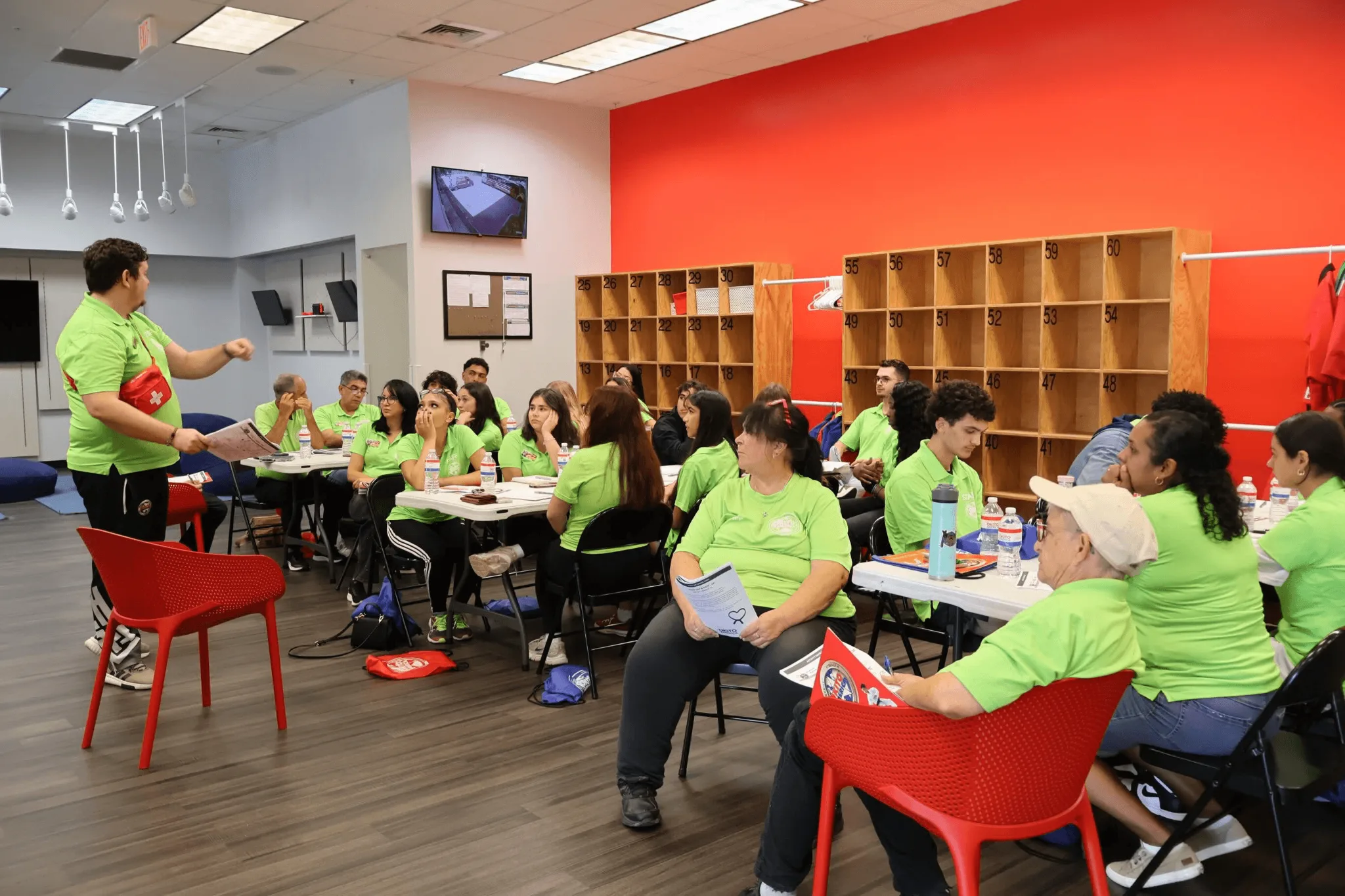 A group of volunteers wearing neon green shirts gather in a modern community center with bright red walls and wooden cubby shelving, seated around tables while a male instructor in a lime green shirt leads a training session.