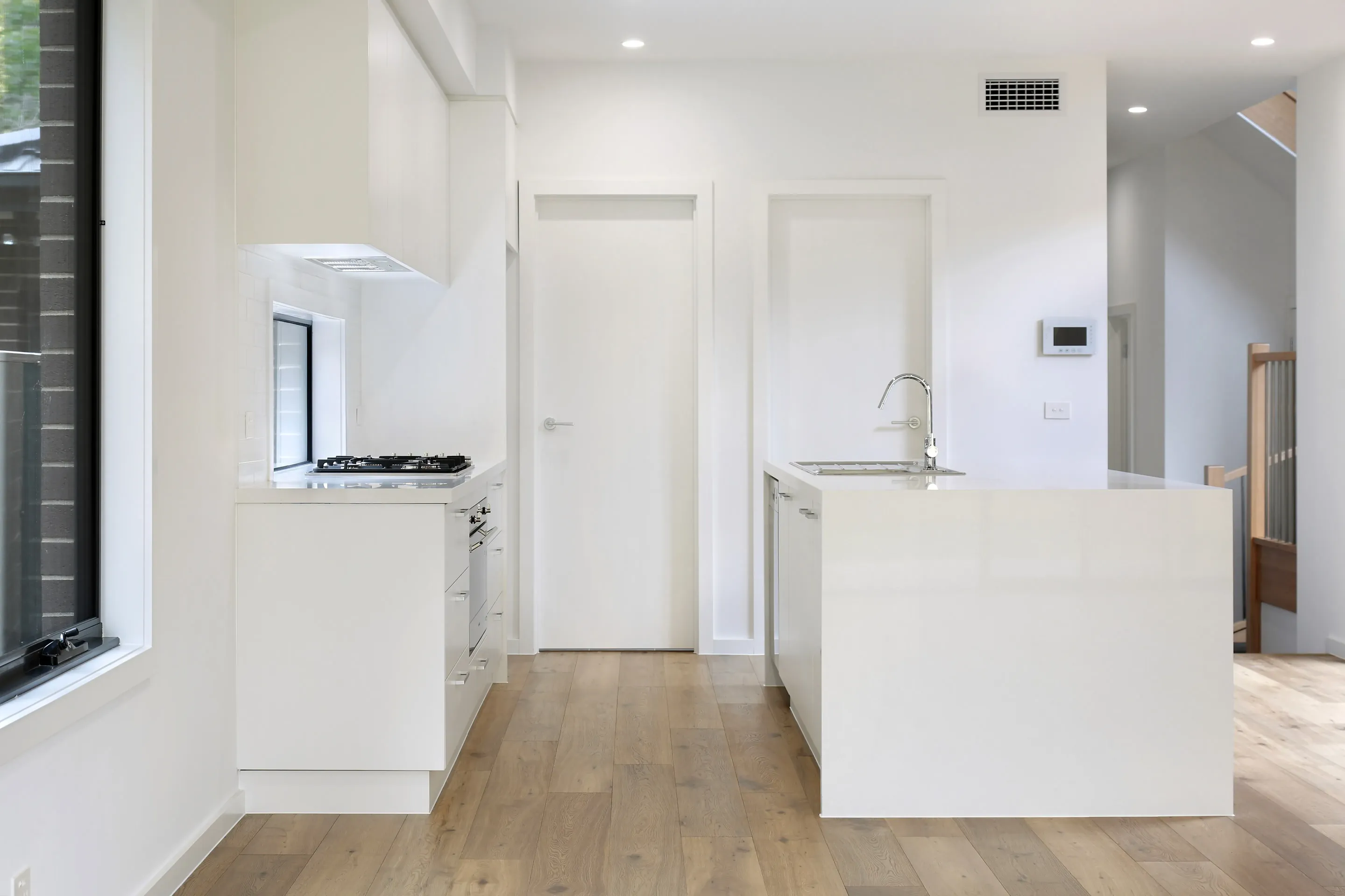 White kitchen with a waterfall island and chrome tap, gas cooktop under a window, white doors to a butler's pantry and a staircase visible beyond, over oak floors