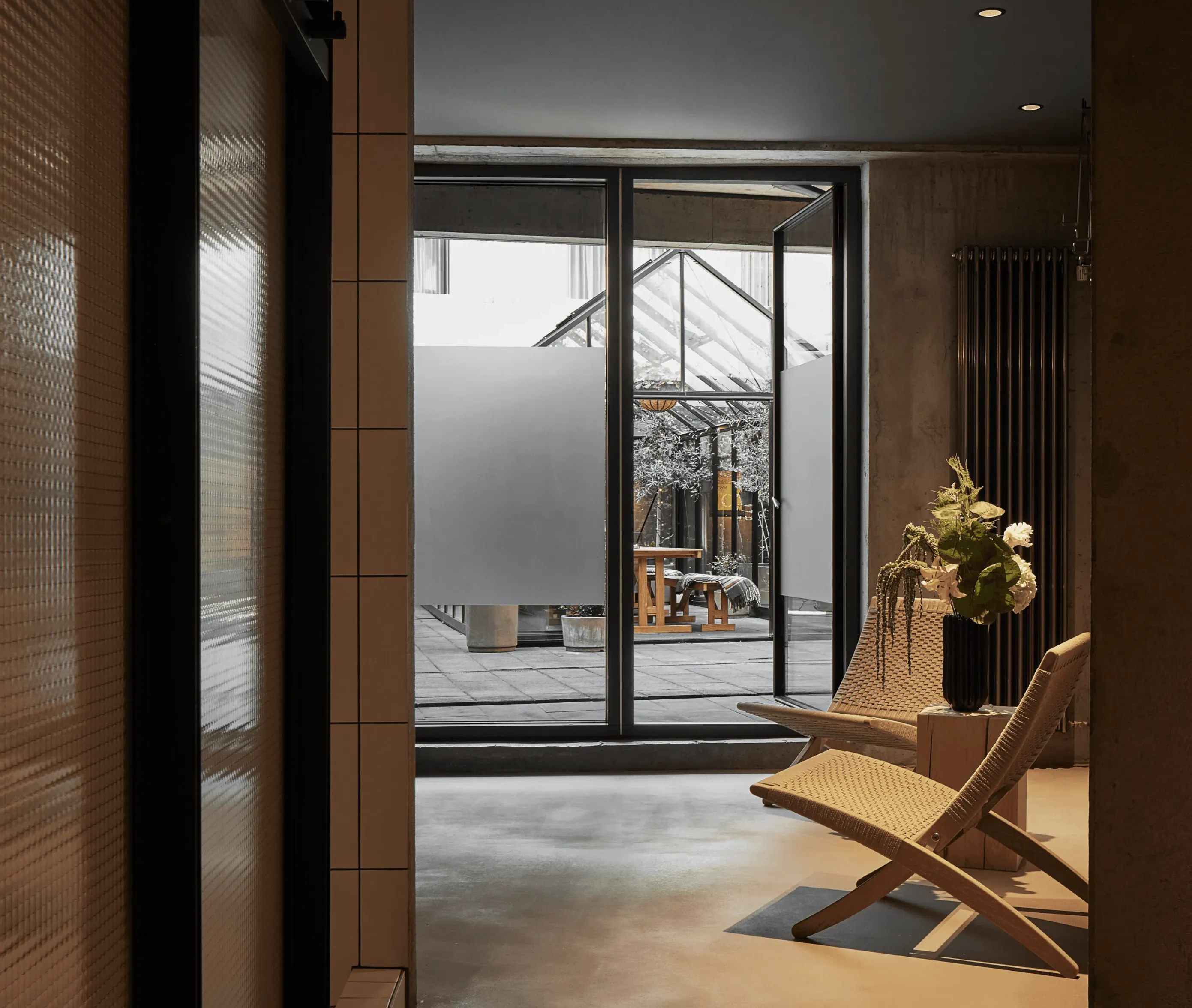 Hotel interior looking through an open glass door toward a bright atrium, with patterned walls, lounge chairs, and a vase of flowers.