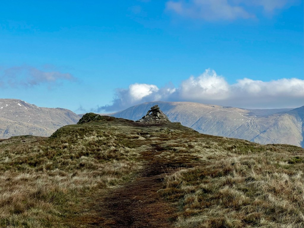 A path leading to the cairn in the distance. Blue skies above and fells in the background.