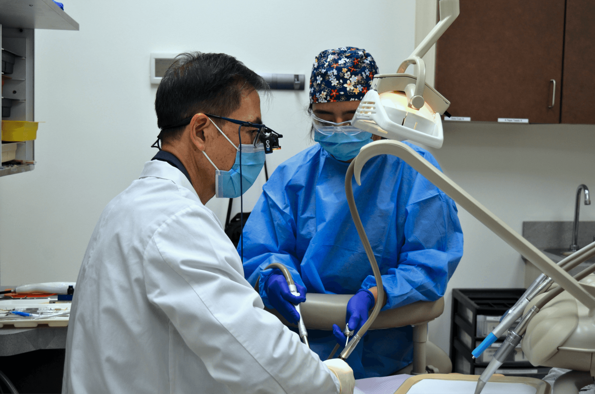 Two dental professionals wearing masks and protective gear performing a dental procedure on a patient in a clinic setting.