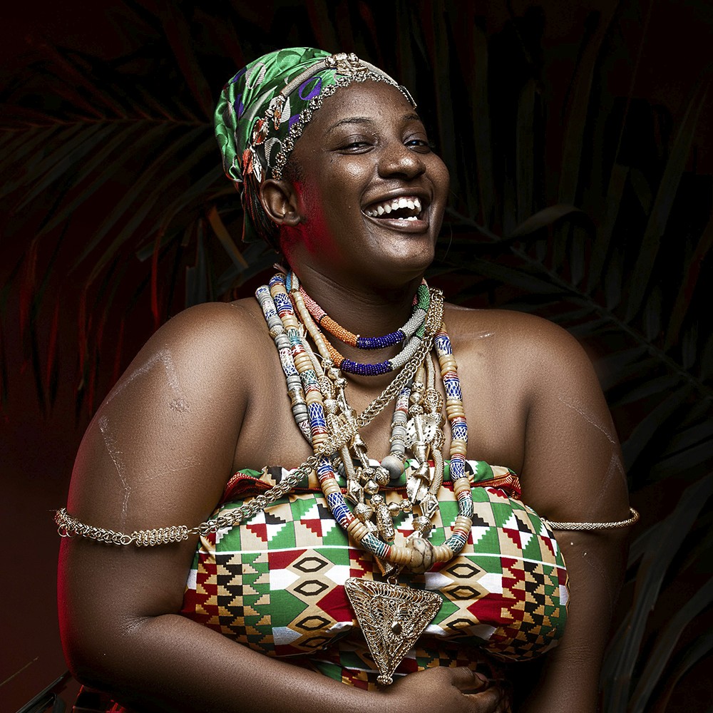 Smiling woman in traditional African attire adorned with colourful beads, jewellery, and a patterned headwrap, standing against a dark background with palm leaves.
