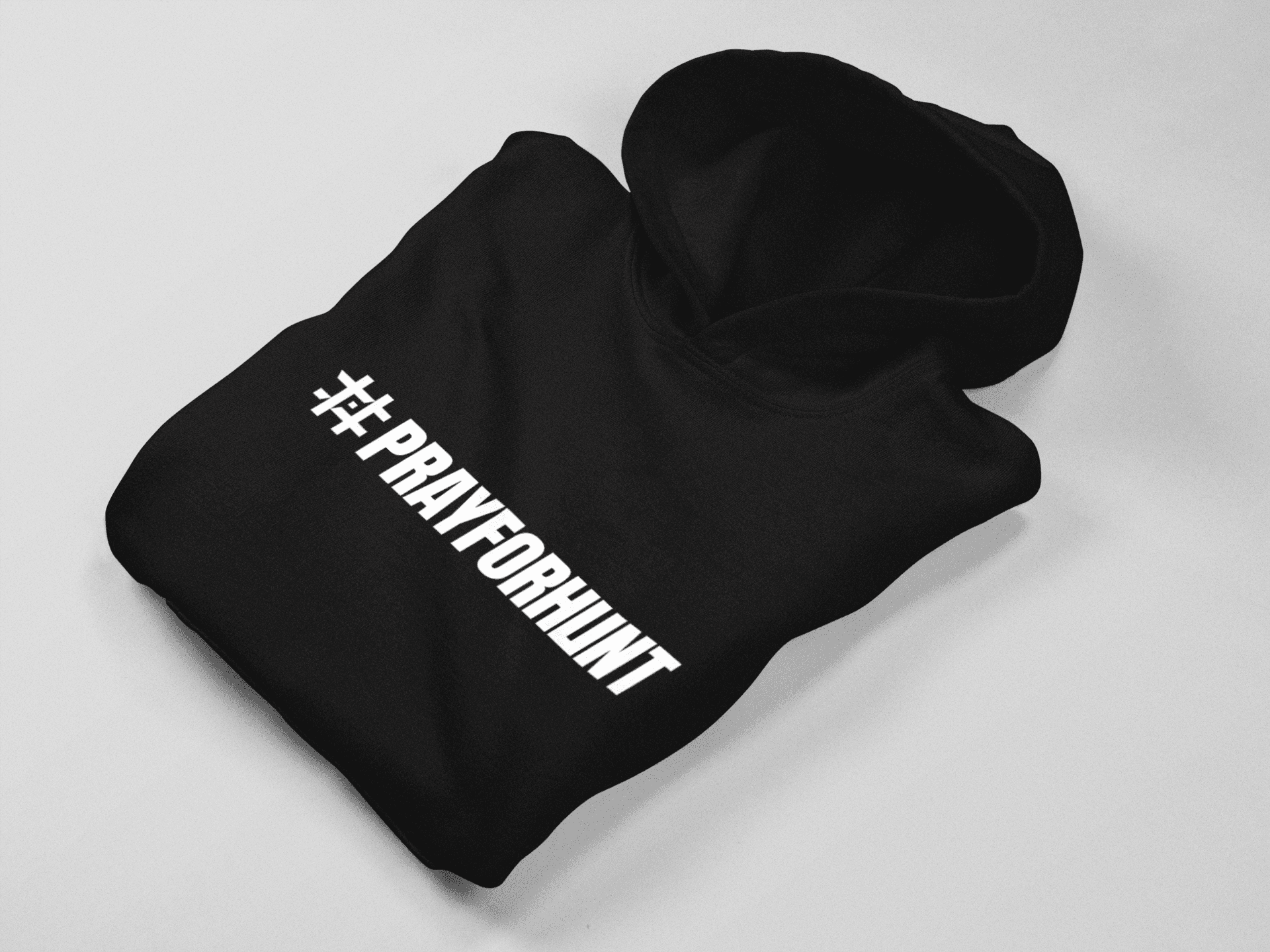 A black hoodie neatly folded on a light, solid surface. The front of the hoodie features the '#PRAYFORHUNT' design in bold, white, all-caps text, with a stylized hashtag symbol at the beginning. The design contrasts sharply with the black fabric, creating a strong and impactful visual. The folded presentation emphasizes the clean and minimalistic style of the apparel.