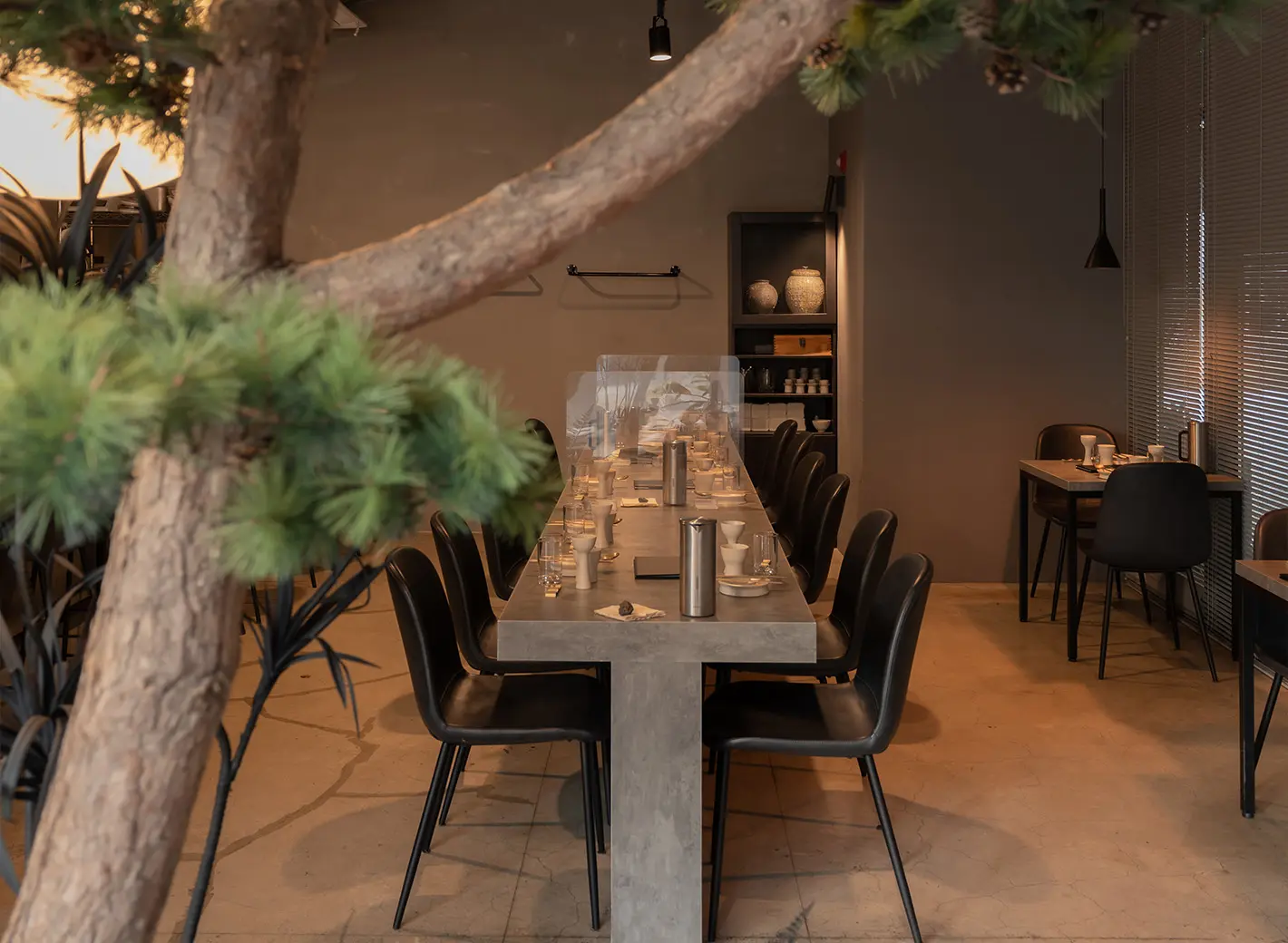 Restauracja Joo052 (주052) – minimalistyczny betonowy stół z czarnymi krzesłami przygotowany do kolacji, a na pierwszym planie gałązka sosny