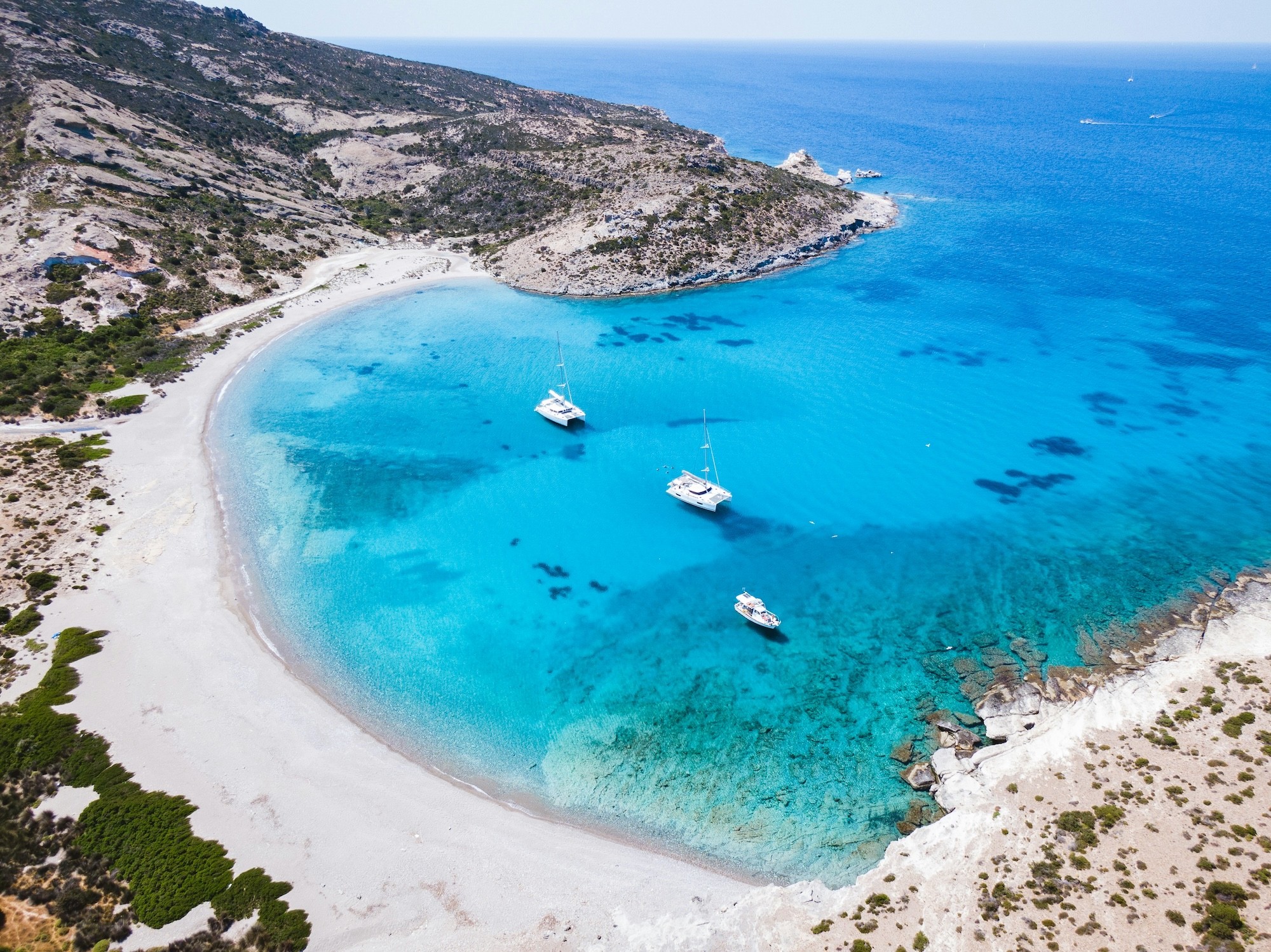 Aerial view of catamarans and sailboats anchored in acrystal-clear bay along a rugged Cycladic island shore
