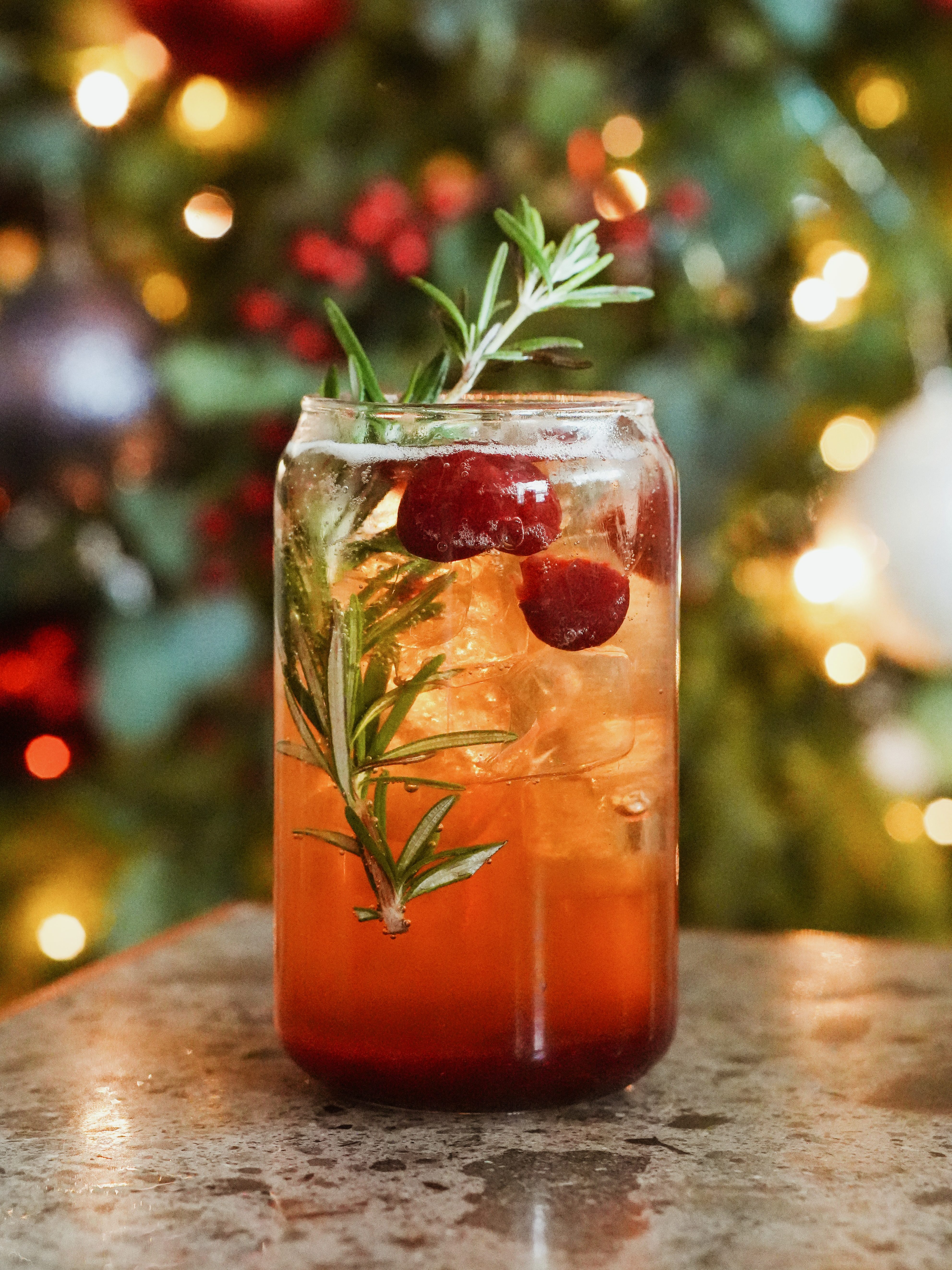 A festive drink garnished with cranberries and rosemary, set against a blurred background of Christmas decorations.