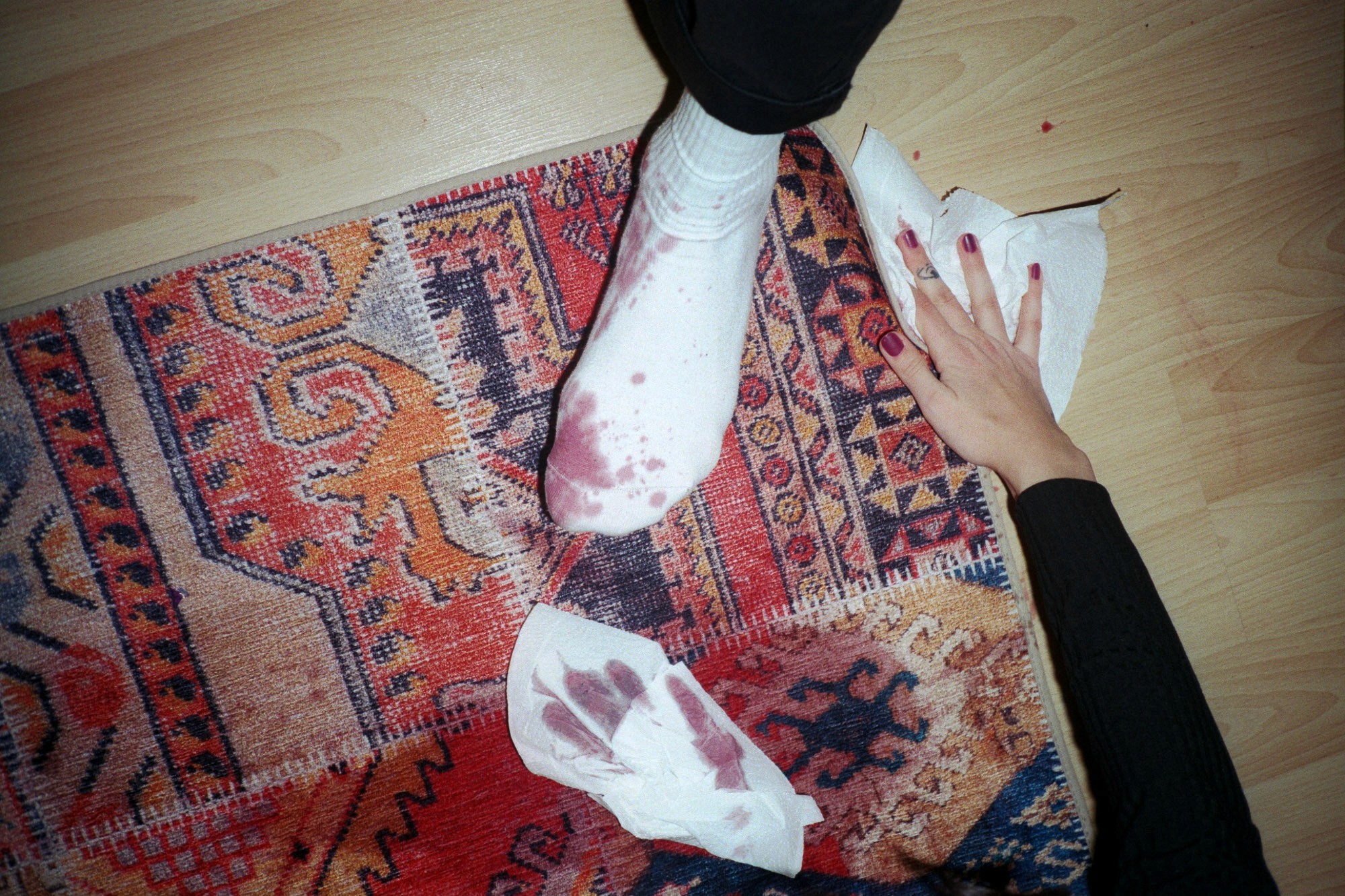 A white sock stained with blood rests on a vibrant, patterned rug, while a hand with painted nails uses a tissue to clean up the bloodstains on the wooden floor.
