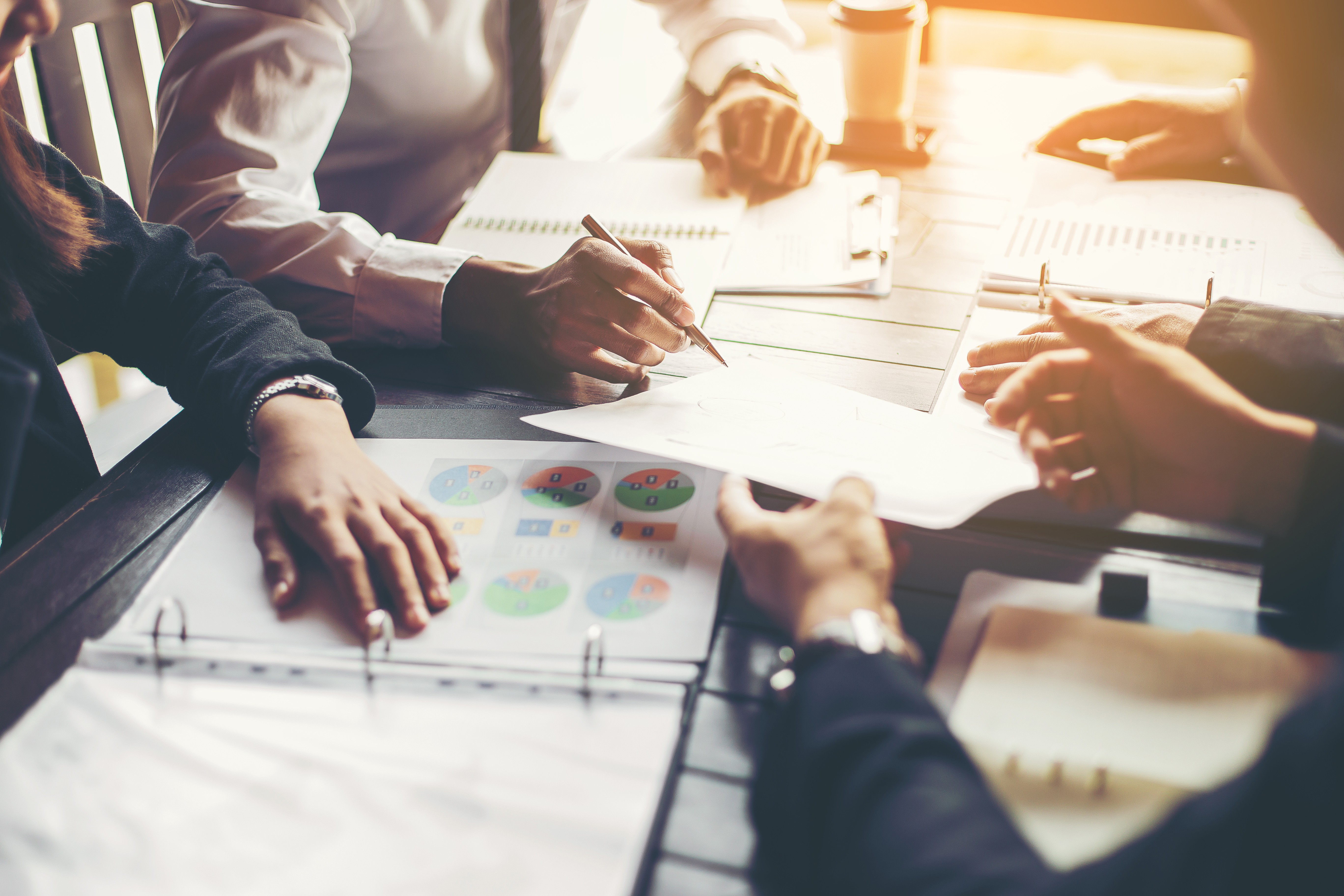Business people around a table reviewing printed charts and documents, with one person writing with a pen