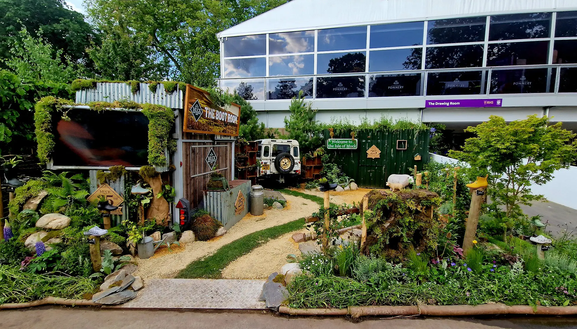 A garden path leads to a modern building, surrounded by greenery and flowers, under a clear blue sky.