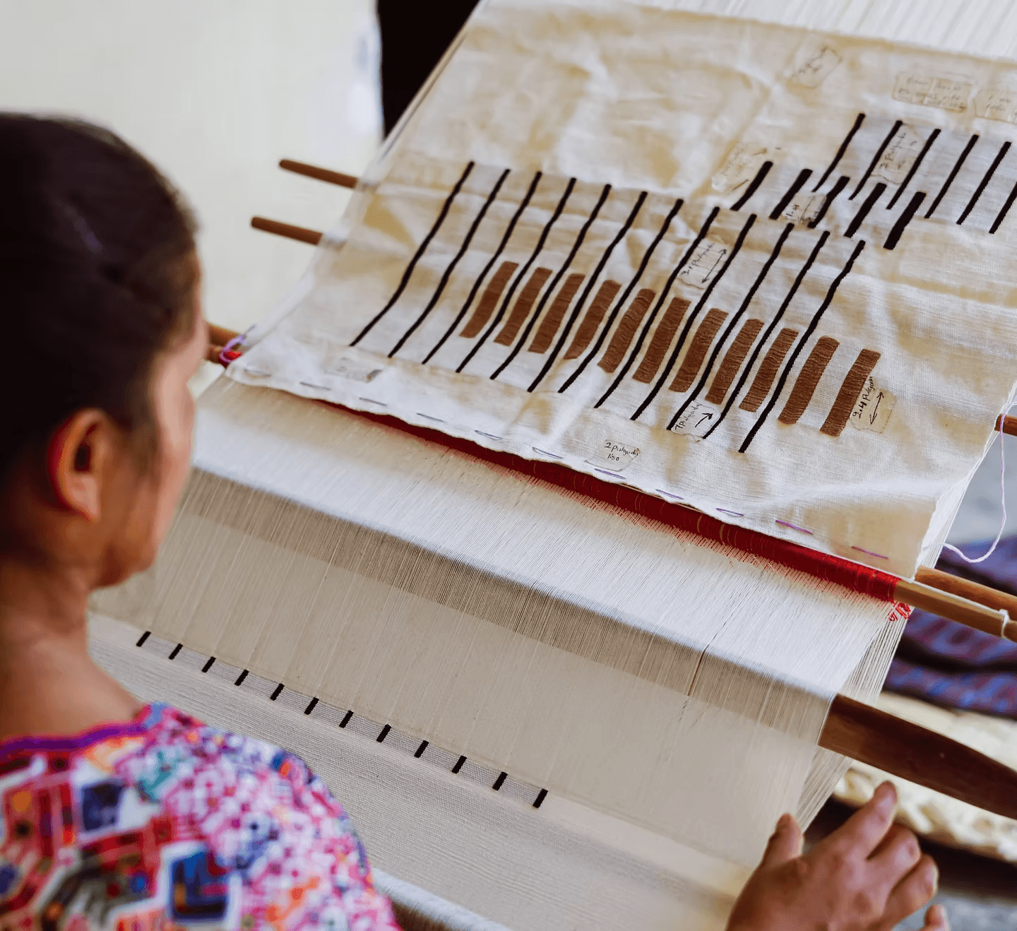 A person wearing a colorful patterned garment is weaving on a traditional loom, with attention focused on intricate black-striped patterns emerging on a white textile.