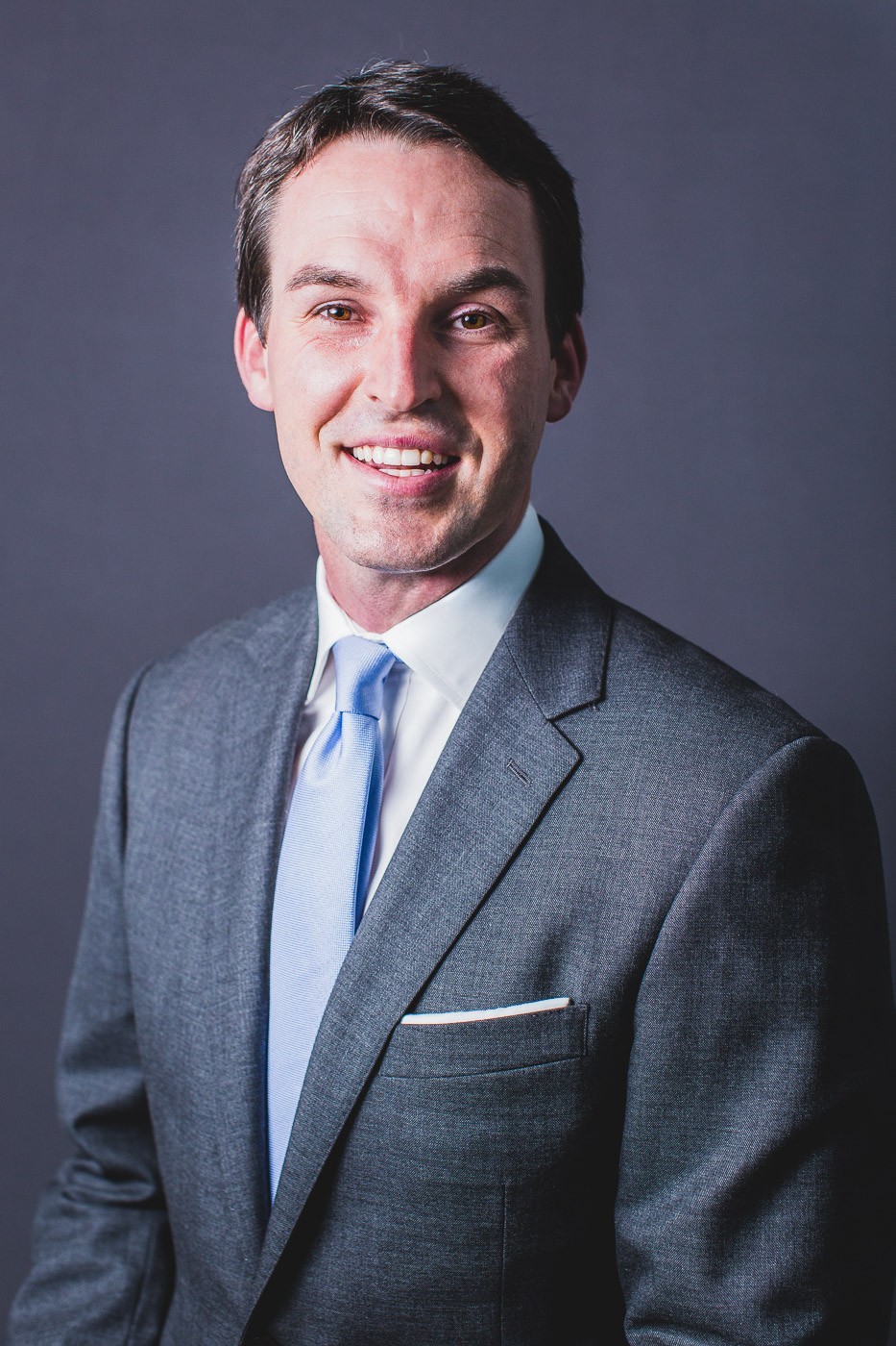 Professional headshot of Jonathan Lusk, Managing Director at Dornick Wealth Management, wearing a gray suit and light blue tie, smiling against a dark background
