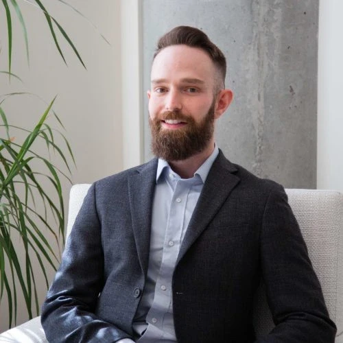 Um homem com uma barba bem cuidada, vestindo um blazer escuro e uma camisa azul-clara, está sentado em uma cadeira branca em uma sala moderna, com uma planta alta e verde e uma coluna de concreto ao fundo.