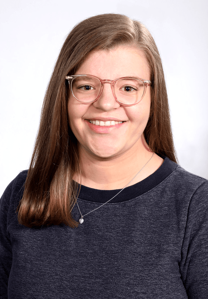 Porträt einer jungen Frau mit langen braunen Haaren und rosafarbener Brille, die lächelnd in die Kamera schaut. Sie trägt ein dunkles Oberteil und eine feine Halskette vor hellem, neutralem Hintergrund. Bei der jungen Frau handelt es sich um Lene Hirschberger