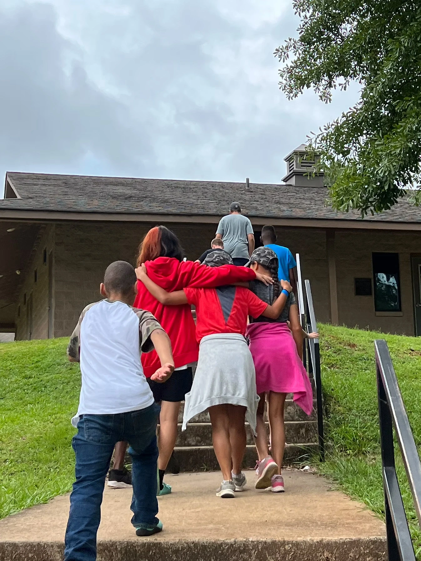 A group of campers ascend a staircase leading to a building. They're walking close together, some with arms around each other.