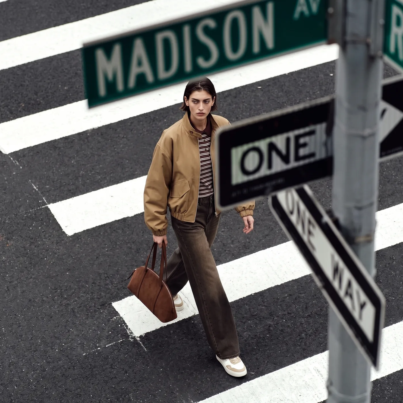 Donna che attraversa le strisce pedonali con bomber cammello, polo a righe, jeans marroni a gamba larga e borsa in suede