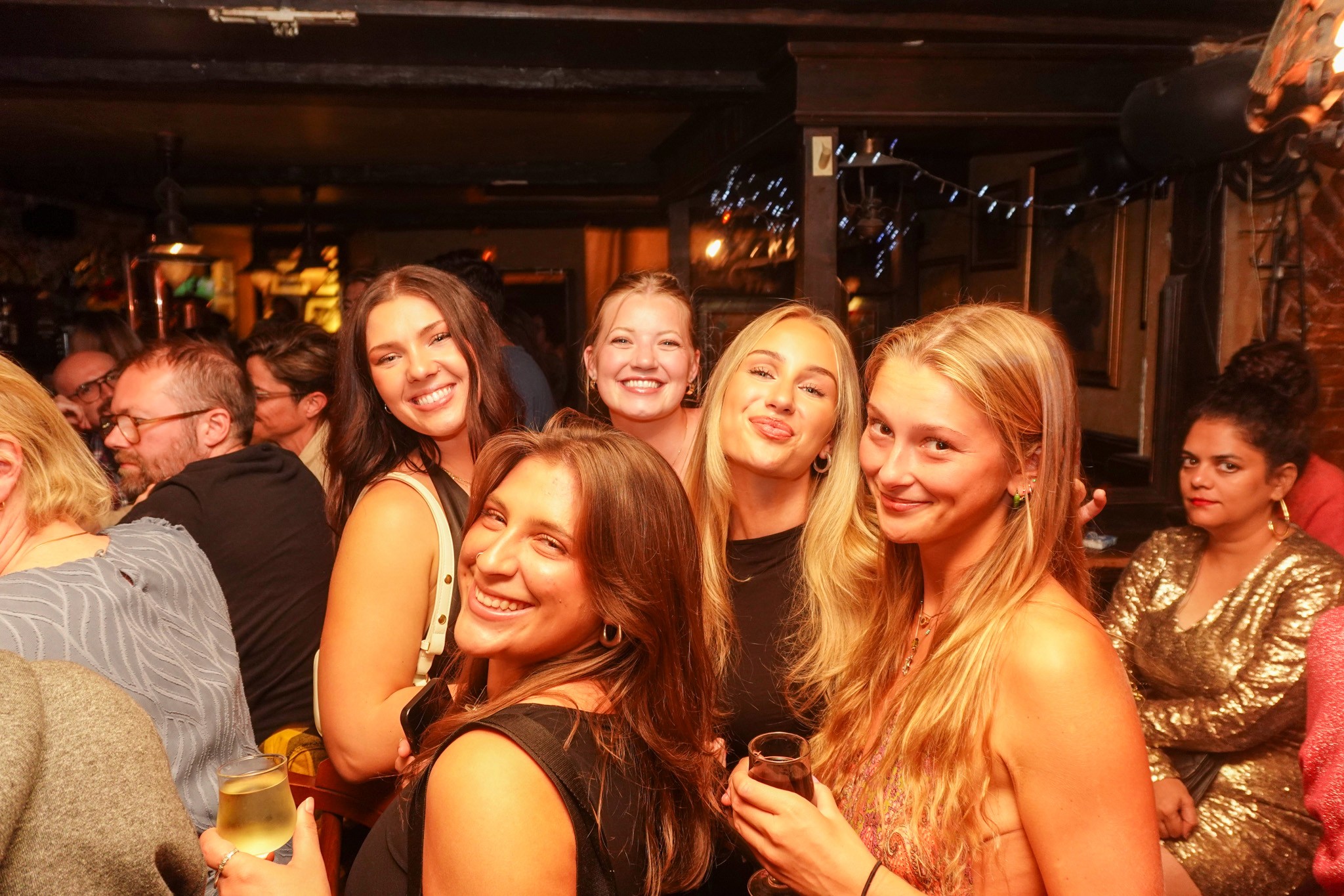Group of young women smiling together inside a lively bar during a bar crawl in Nice holding drinks and posing under warm lights capturing friendship nightlife energy and the social atmosphere of a French Riviera party experience
