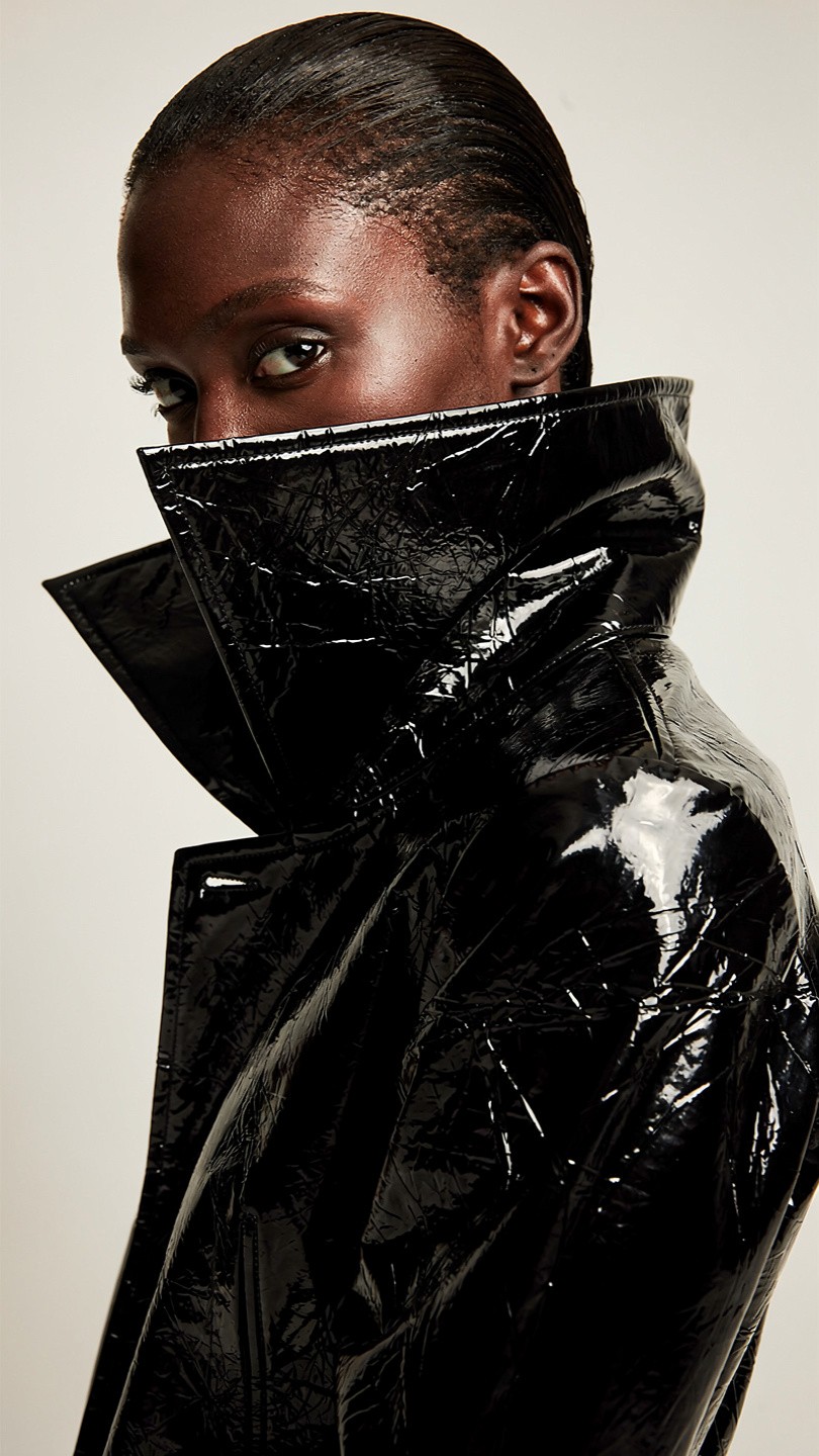 A person wears a shiny black vinyl coat with a high collar, creating a dramatic and modern look. The expression is confident and bold against a neutral background.