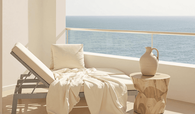 Sunlit balcony with cushioned lounger, wooden side table, and ocean view.