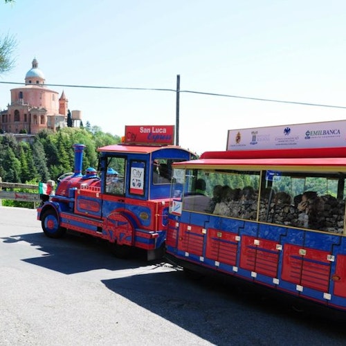 Un piccolo trenino turistico blu e rosso chiamato "San Luca Express" è parcheggiato su una strada con una vista lontana della basilica in cima alla collina.