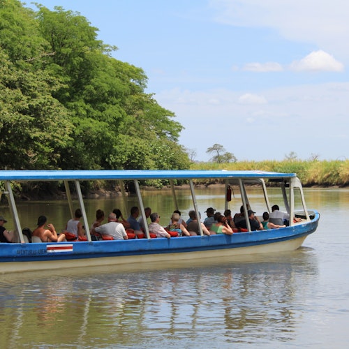 เรือทัวร์หลังคาสีฟ้าที่เต็มไปด้วยผู้คนแล่นไปตามแม่น้ำที่สงบข้างต้นไม้เขียวและตลิ่งหญ้าภายใต้ท้องฟ้าที่มีเมฆบ้างเล็กน้อย