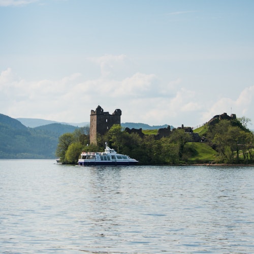 一艘游船在一个宁静湖泊中的一座郁郁葱葱的小岛附近巡游，岛上有一座古老、部分损毁的石头城堡，远处是连绵的群山。