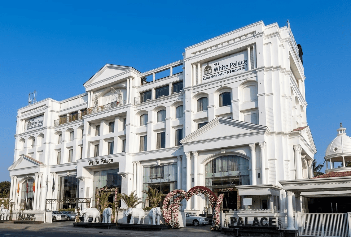 Two-story white house with a wraparound balcony overlooks a clear blue swimming pool. Palm trees and potted plants add a tropical vibe under a sunny sky.