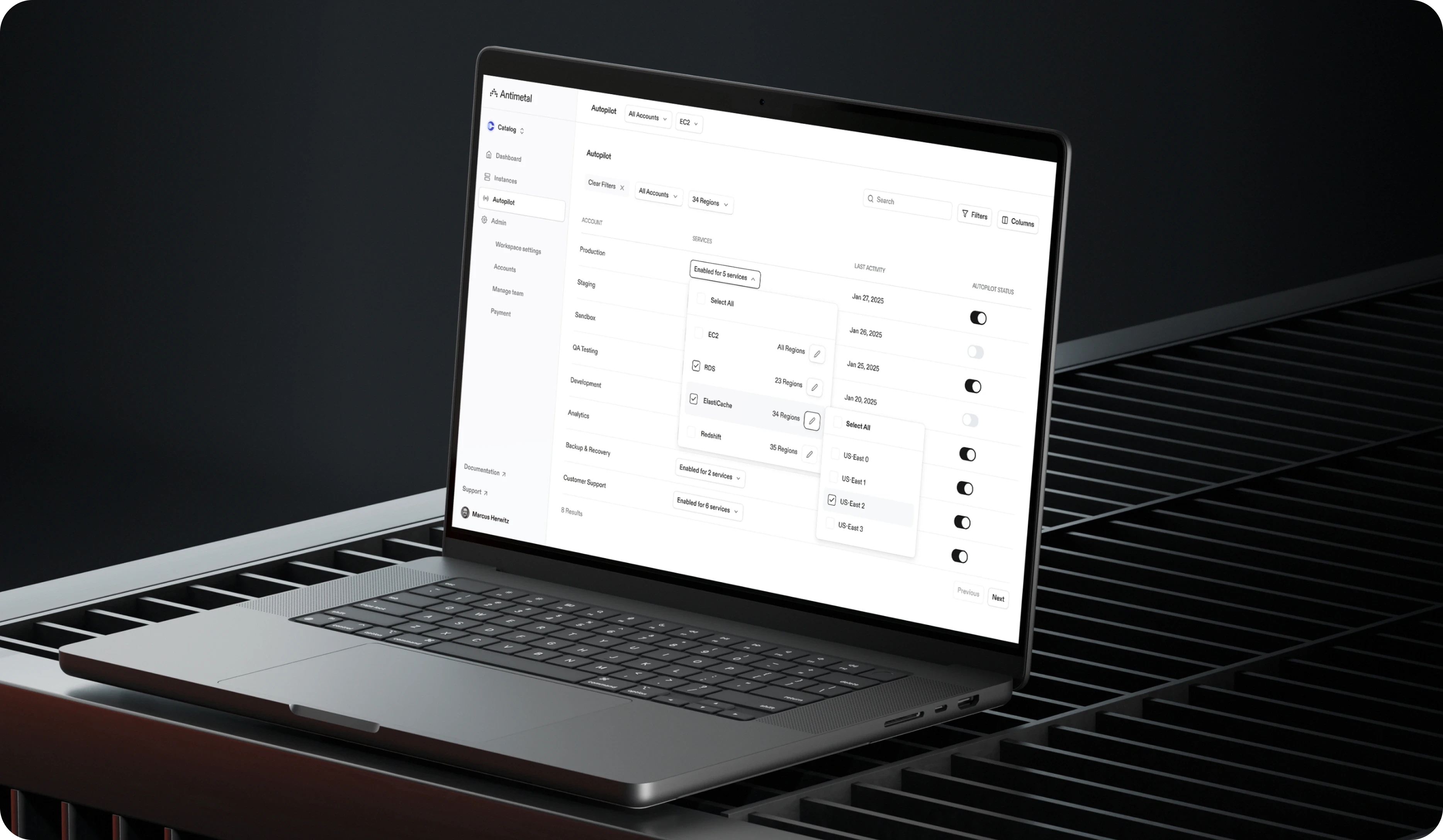 Laptop displaying Antimetal's "Autopilot" feature interface, showing a dashboard with toggles for enabling services across different regions and accounts. The laptop is placed on a sleek, dark metal surface with a grid pattern, against a minimalist black background, emphasizing a professional and modern design aesthetic.