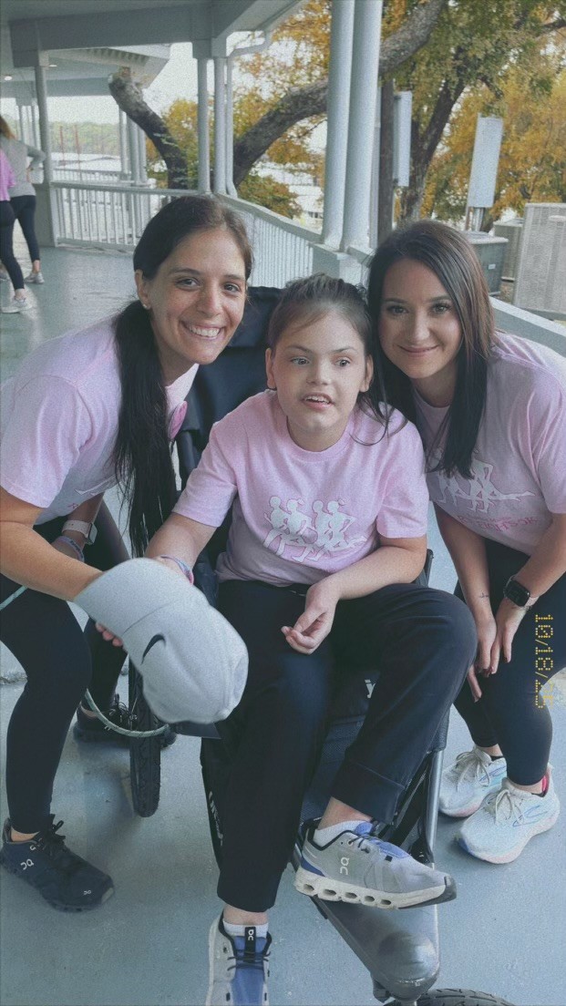 A smiling young girl in a wheelchair is flanked by two women wearing matching pink shirts and black pants, posing together on a covered porch with trees and a railing in the background.