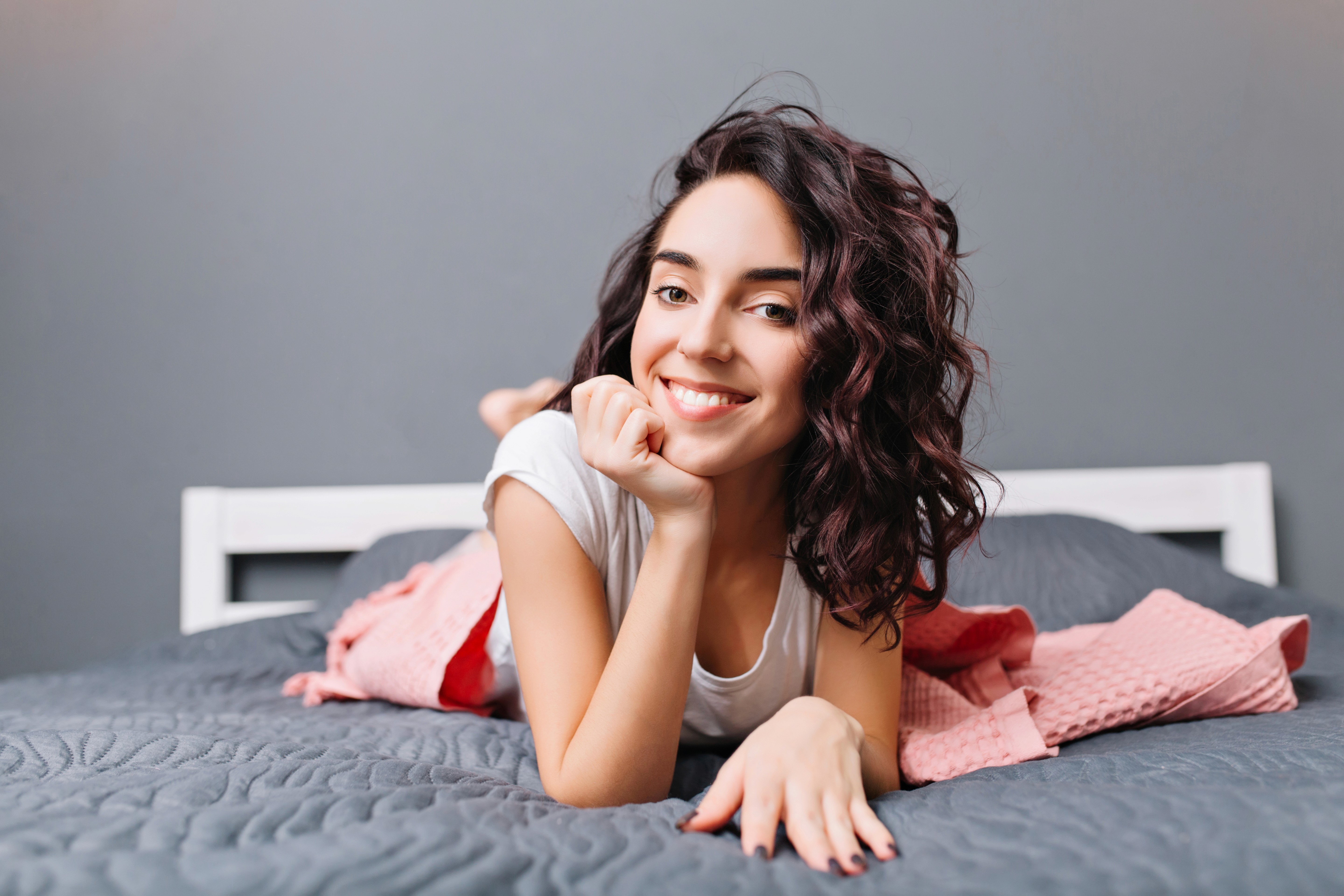 Smiling woman with curly hair lies on a gray bedspread, wearing a white shirt. A pink blanket is beside her, creating a cozy, cheerful atmosphere.