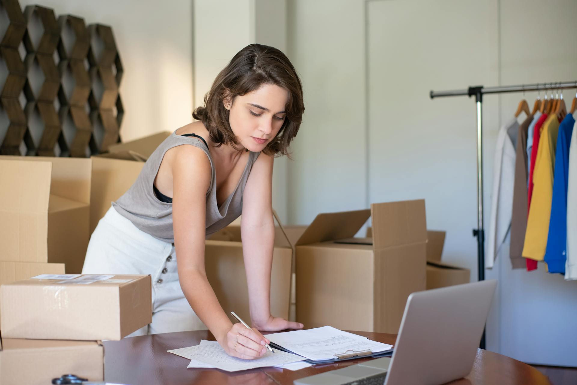 A young woman with dark hair, wearing a gray tank top and white skirt, is leaning over a wooden table to write on a document or checklist. She is surrounded by stacked cardboard boxes, suggesting she is preparing for a move or managing an e-commerce business. A laptop and a rack of colorful clothes on hangers are also visible in the background.