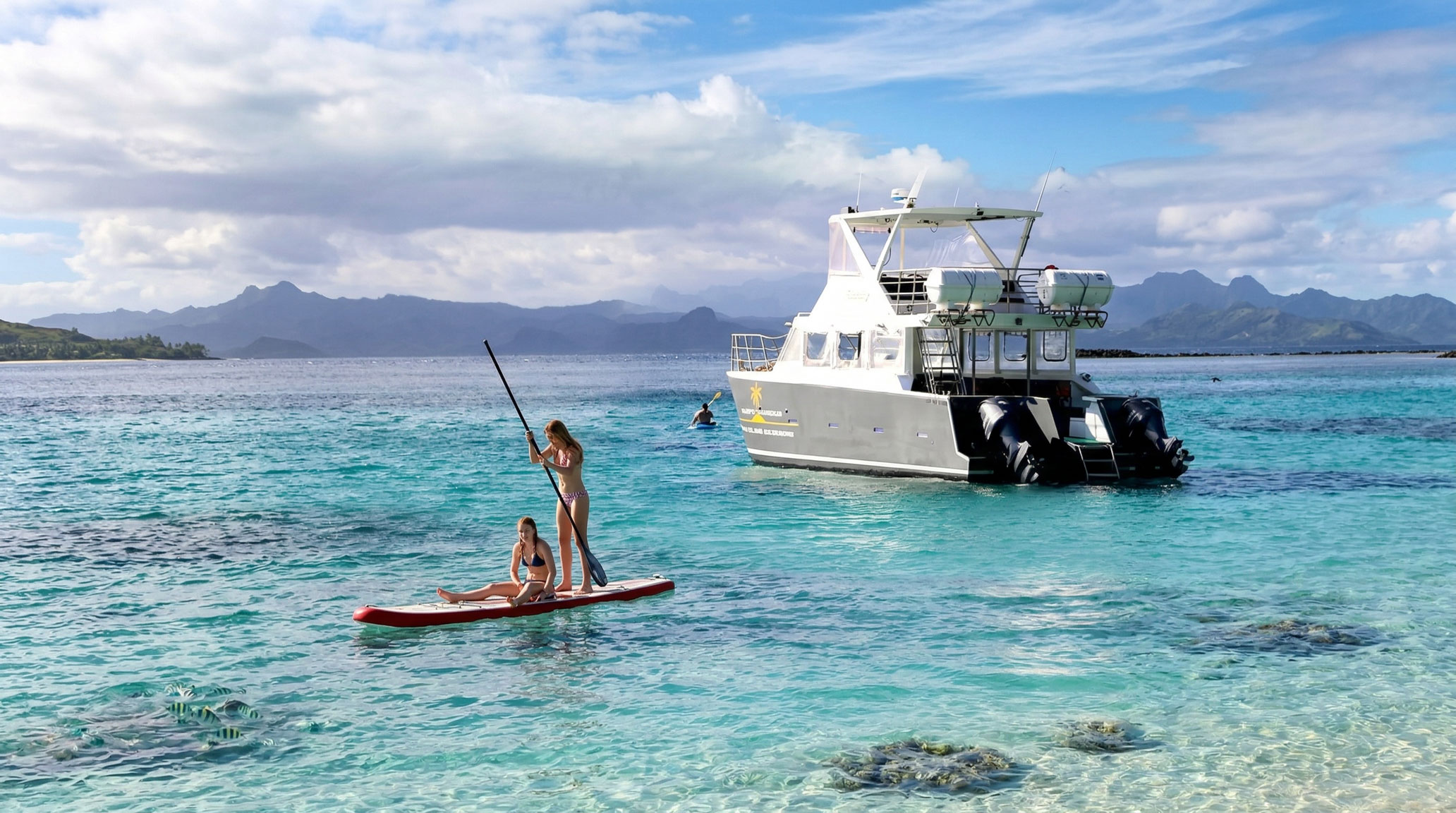 Two women paddle board in clear turquoise waters with a tour boat and distant mountains in Fiji