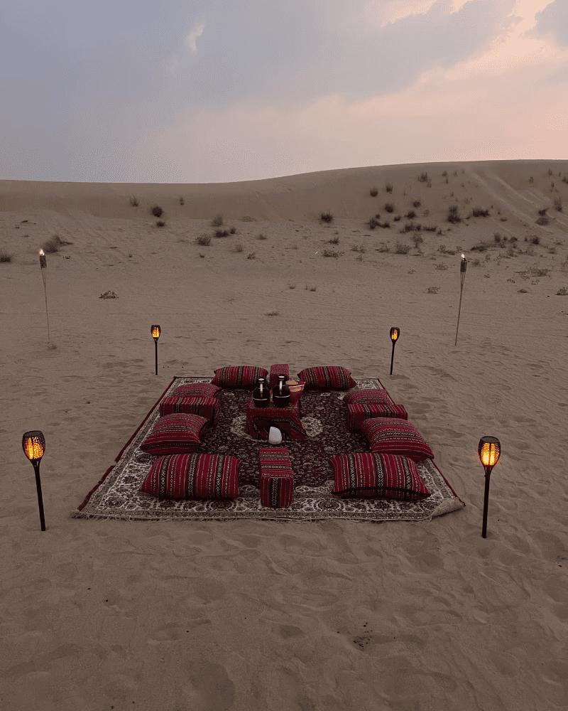 Canvas tent set up on sandy desert dunes at sunset, with the sun setting behind the horizon and a clear sky above.