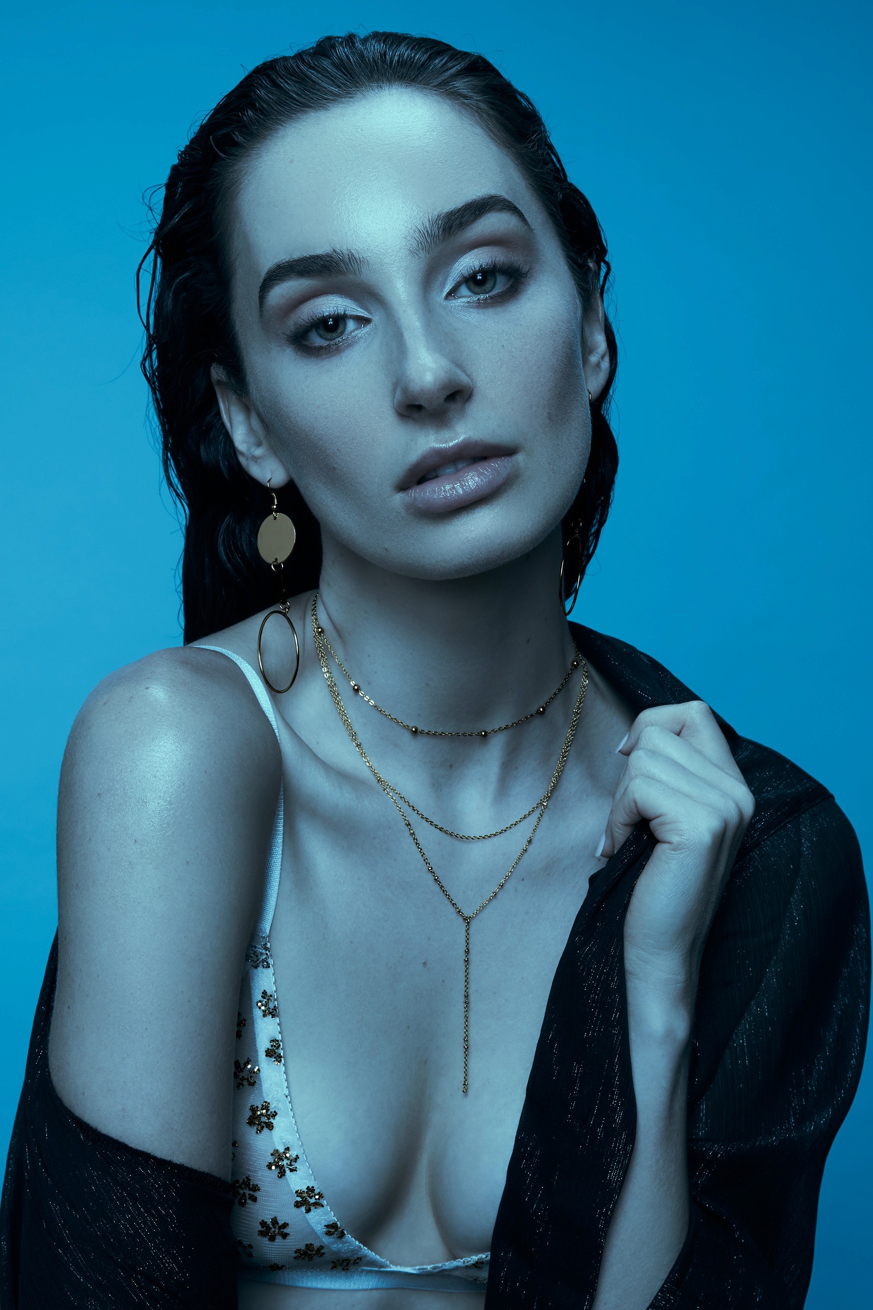 Fashion photography of a female brunette model in soft cool lighting wearing a white bralette with gold details, and a sheer black shirt off one shoulder.