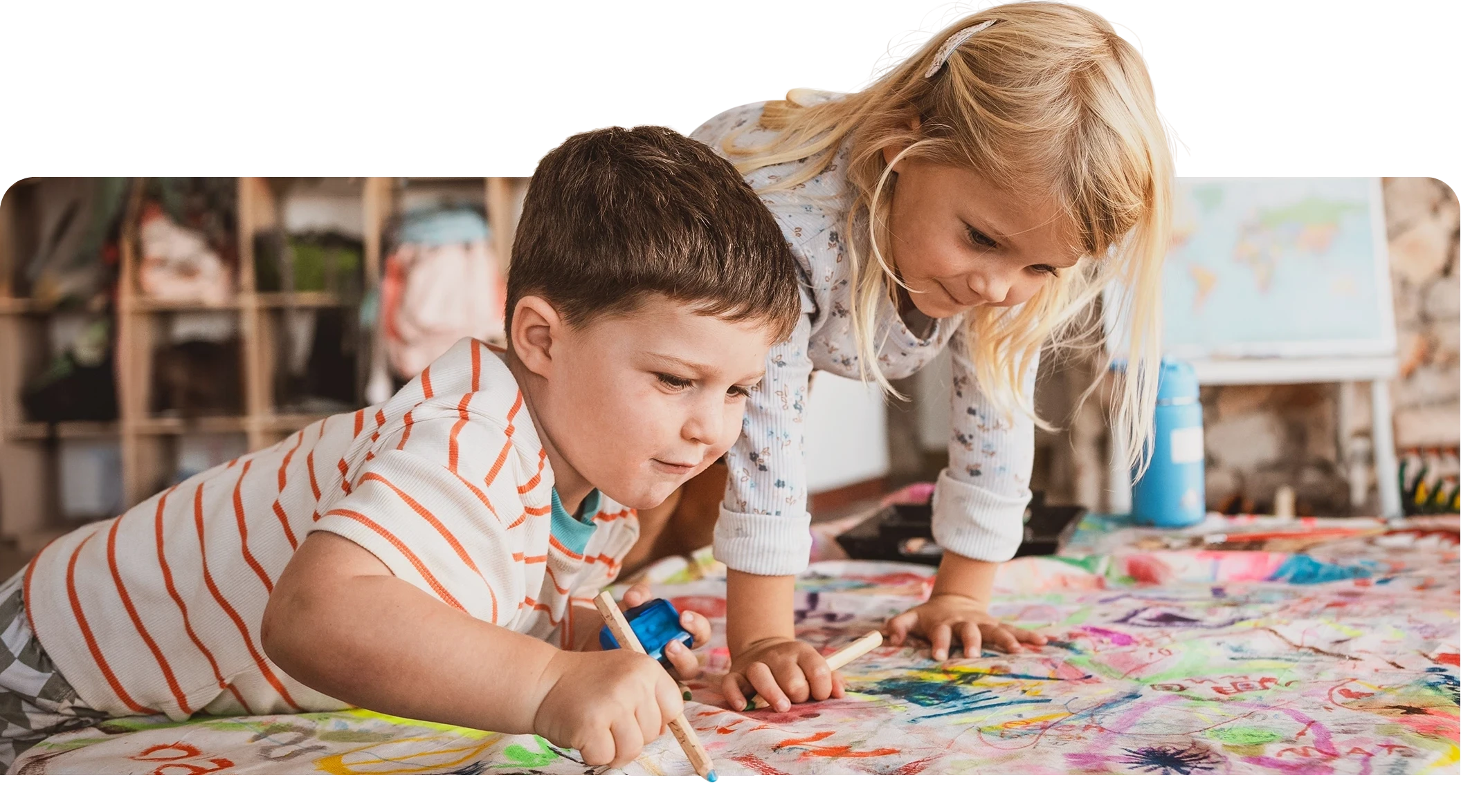 Two young children in a classroom lean over a table covered in colorful drawings, focused on coloring together.