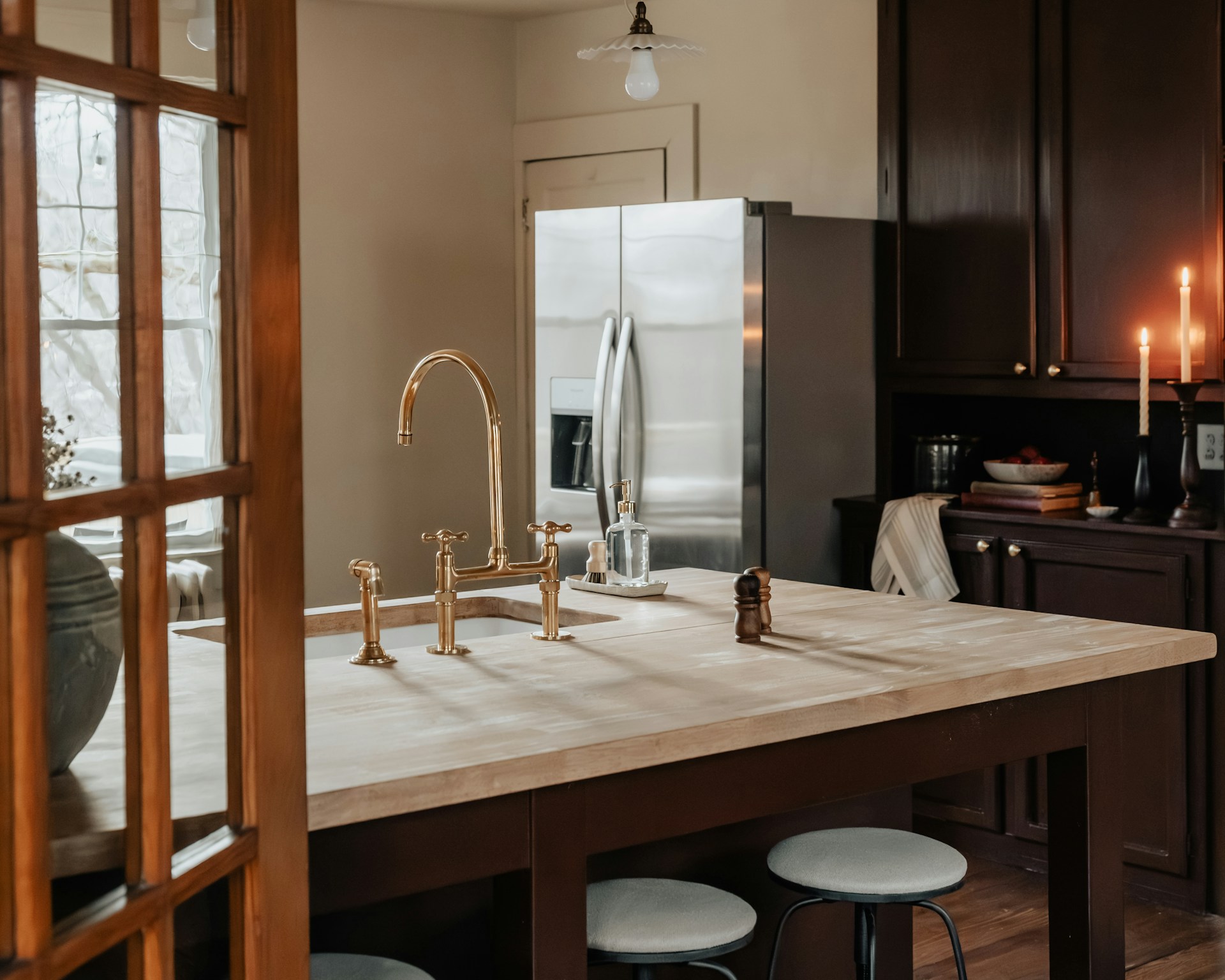 Warm kitchen with a gold faucet on a wood island, stools, dark cabinets, lit candles, and a stainless steel refrigerator.