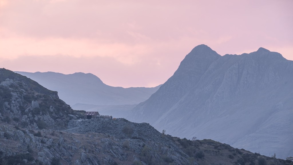 A small slate mine on a hill in front of the jagged Pike of Stickle mountain range. A purple haze across the image.