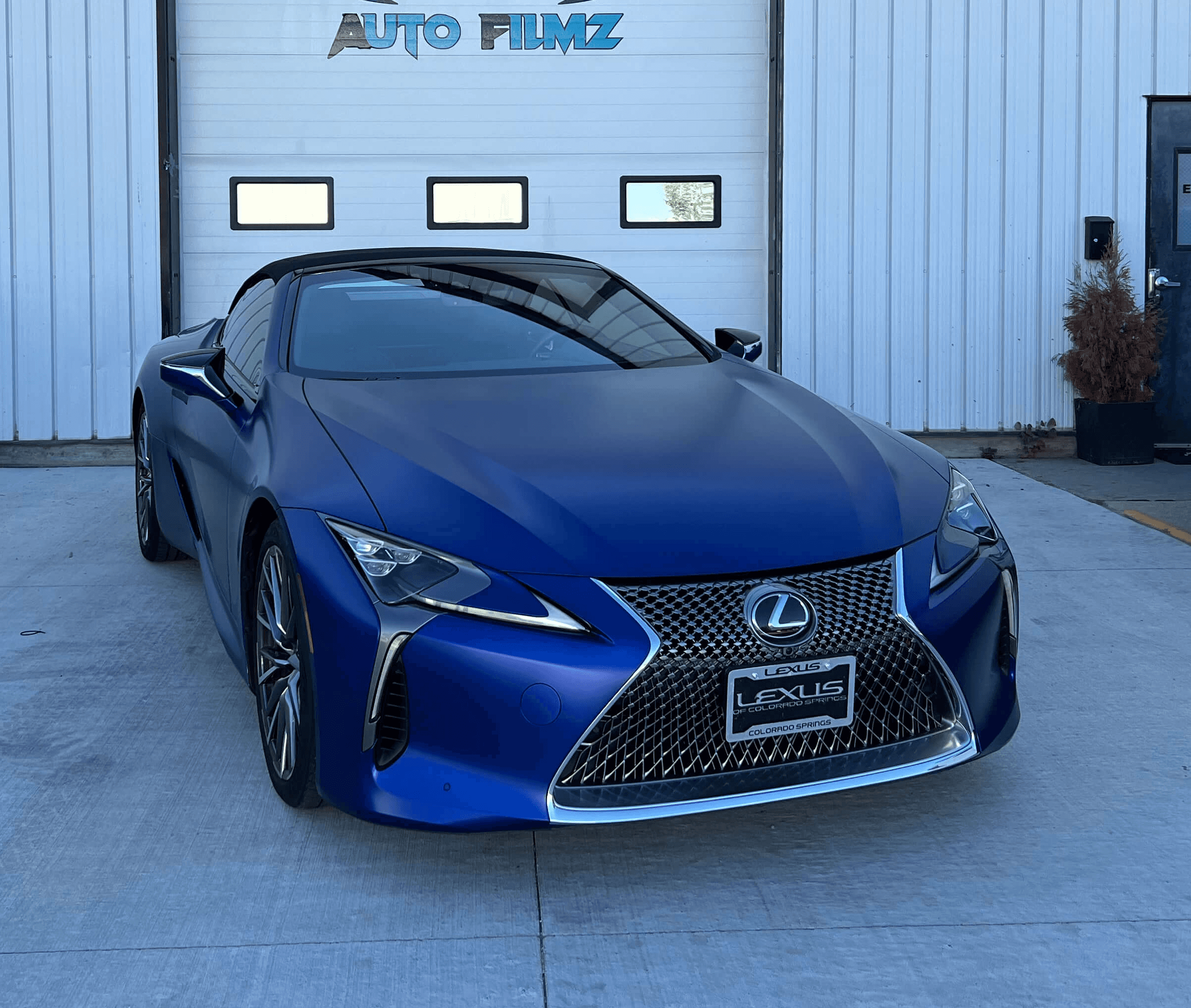 A striking blue Lexus LC coupe is parked on a concrete driveway in front of a white industrial building.