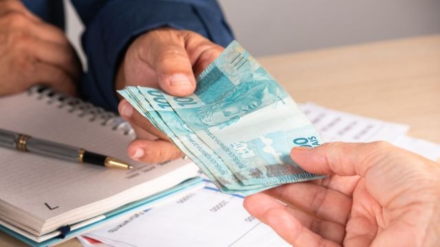 Pessoa entrega cédulas de real a outra pessoa sobre mesa com caderno e documentos